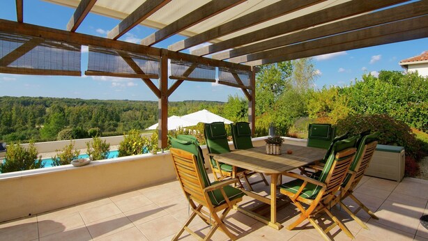 An outdoor patio with a wooden table and chairs, a view of a swimming pool, and a covered area with a pergola.