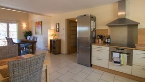A modern kitchen with a stainless steel refrigerator, oven, and a built-in range hood. There is a dining area with wicker furniture and a small table with a vase.