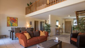 A spacious living area with a high ceiling, a wooden balcony, a large sofa, a coffee table, and a dining table.