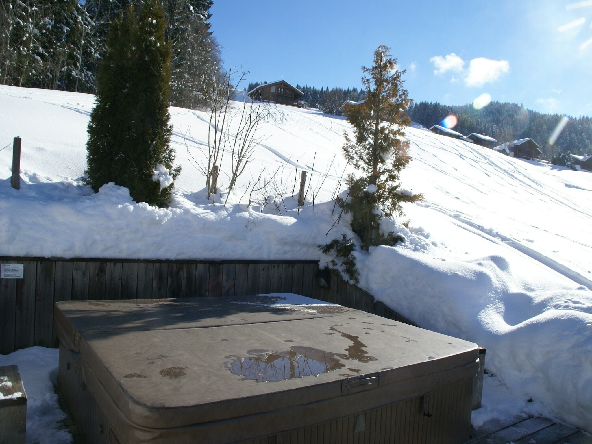 Una terraza de madera con un jacuzzi en un área exterior nevada, rodeada de árboles y una casa al fondo.