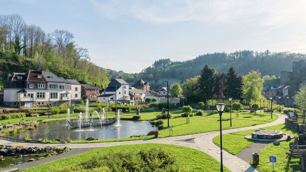 A riverside park with a fountain, pathways, and buildings in the background.