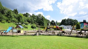 A playground with various play structures including slides and climbing areas, surrounded by a grassy field and trees.