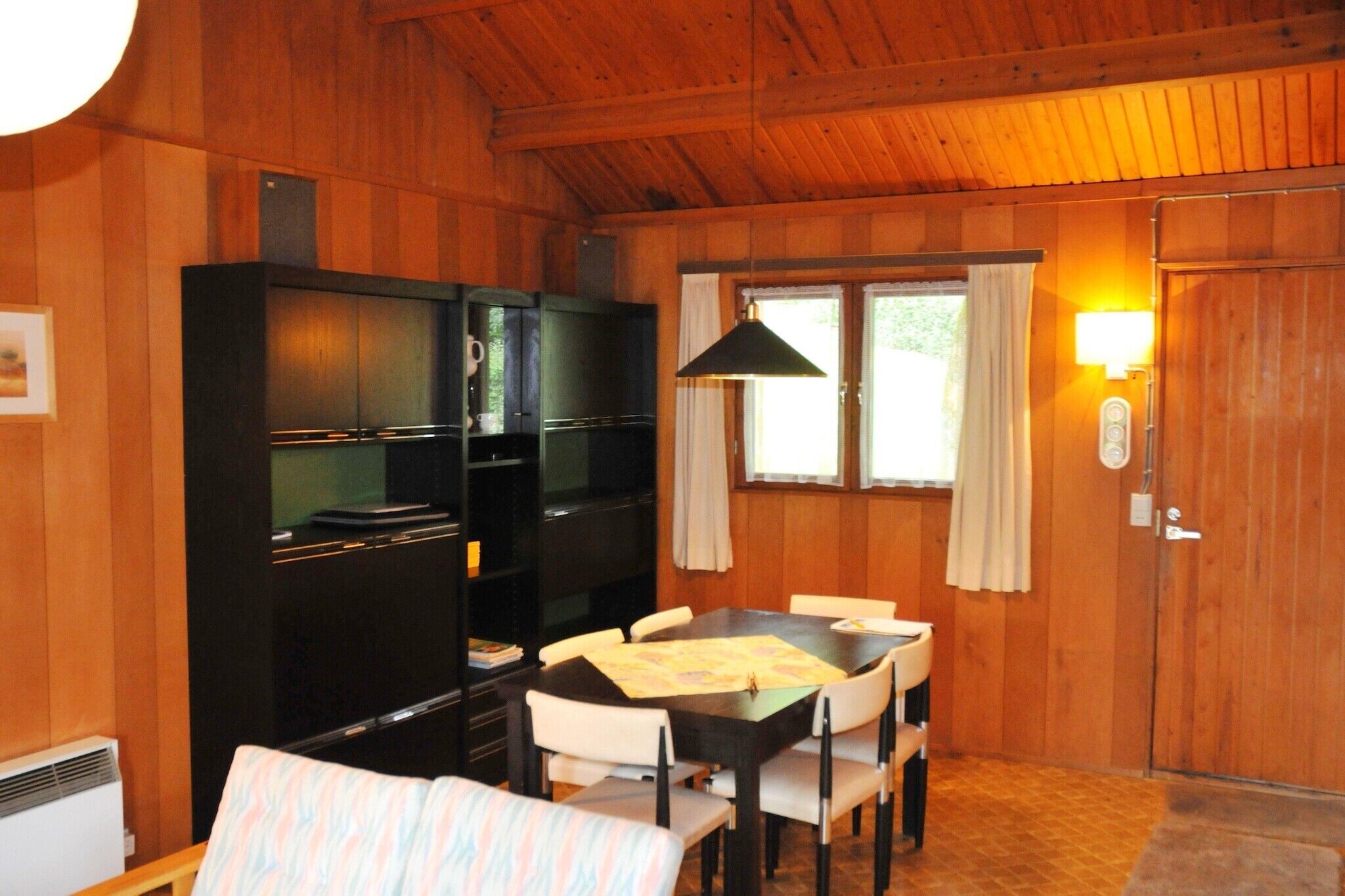 A wooden-paneled room with a dining table, chairs, a black cabinet, and a window with curtains.