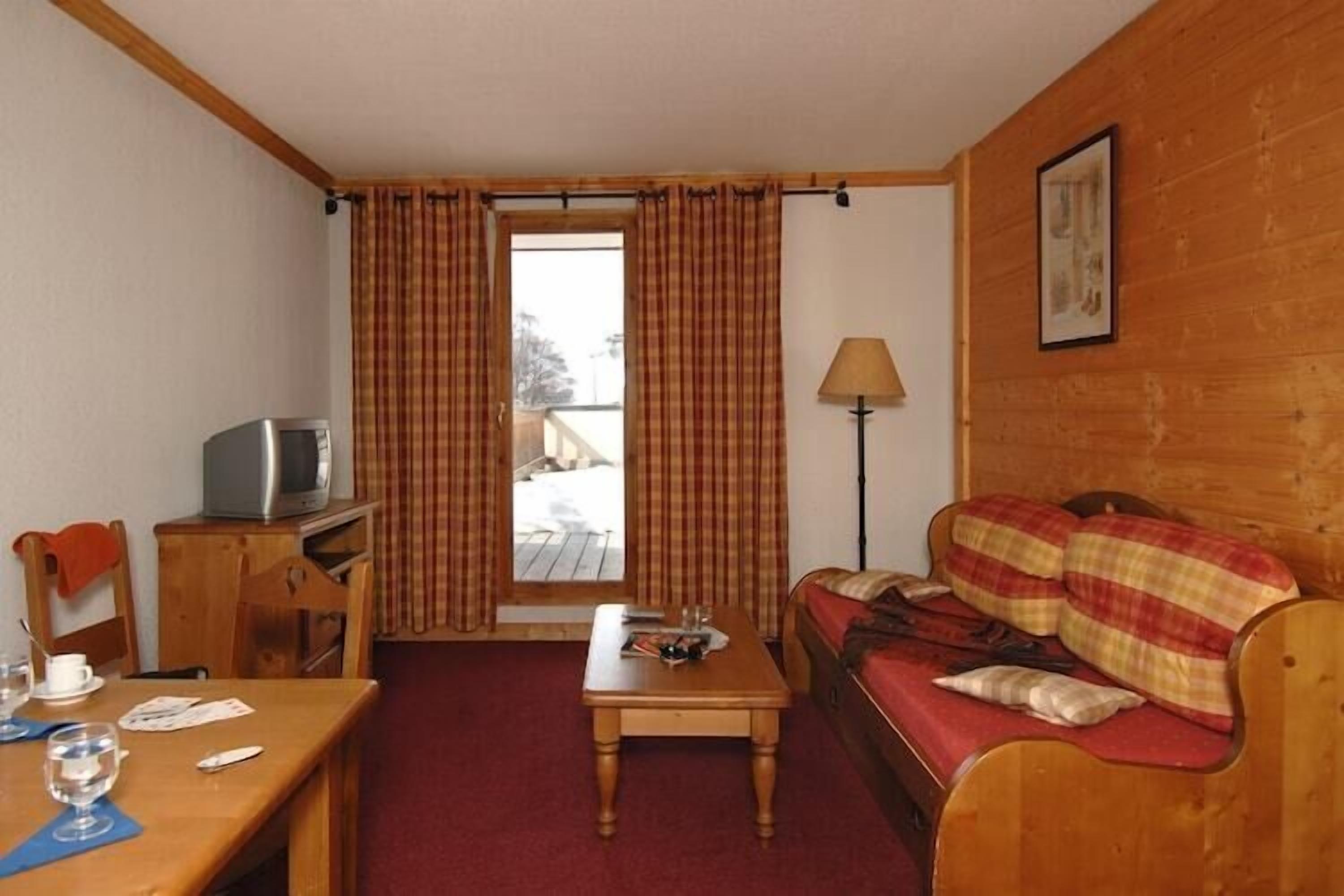 A room with a wooden sofa, a small table, a television on a wooden stand, and a window with curtains.