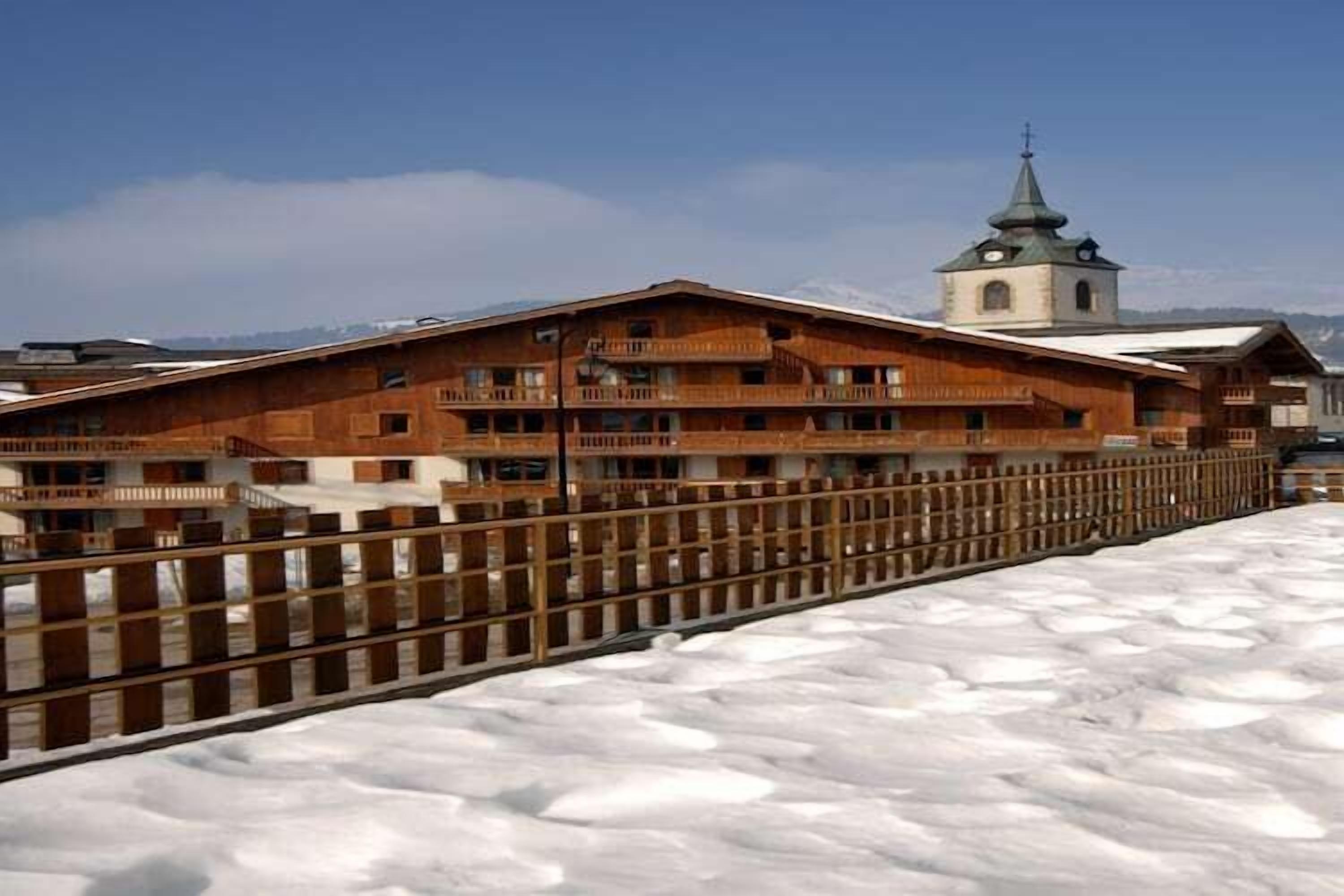 Un edificio de madera con balcones, suelo cubierto de nieve e iglesia al fondo.