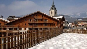A wooden building with balconies, a church spire, and a snow-covered ground. - Apartment in Le Village with Balcony (Notre Dame de Bellecombe)
