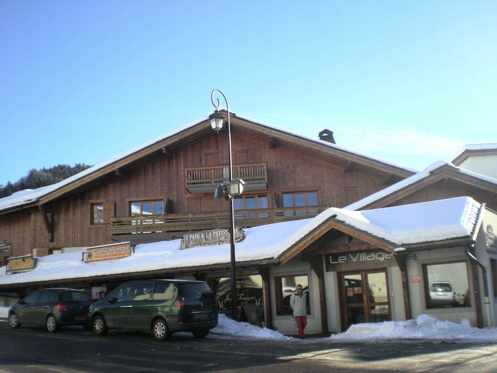 A wooden building with a sign for 'Le Village' and a sign for 'Le Pain & La Patissie.' There are cars parked in front and a person standing in front of the building.