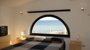 A bedroom with a checkered bedspread, a TV on a wooden cabinet, a lamp, a bedside table, and a window with a view of the sea.