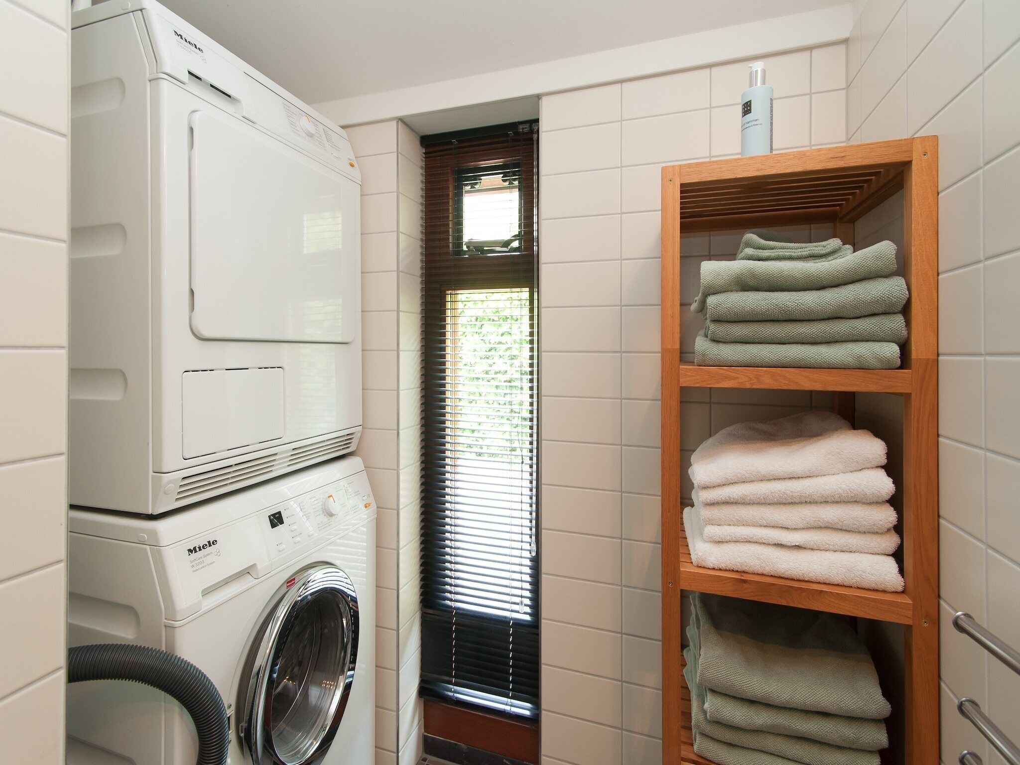 A laundry room with a front-loading washing machine and dryer, a wooden shelf with folded towels, and a window with blinds.