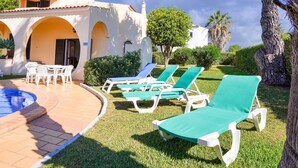 A pool area with lounge chairs and a table, surrounded by greenery and a building with arches.