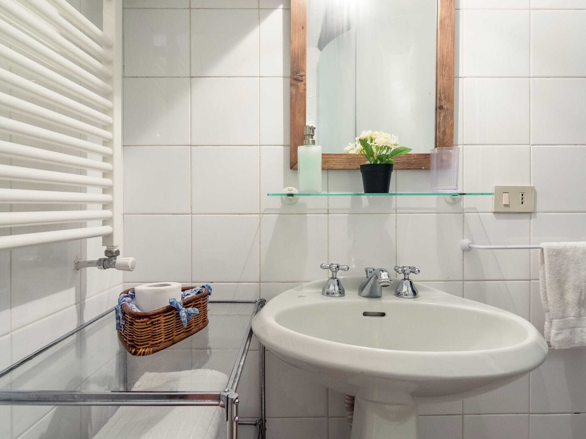 A bathroom with a white sink, two faucets, a glass shelf with toiletries, a wicker basket, and a towel rack.