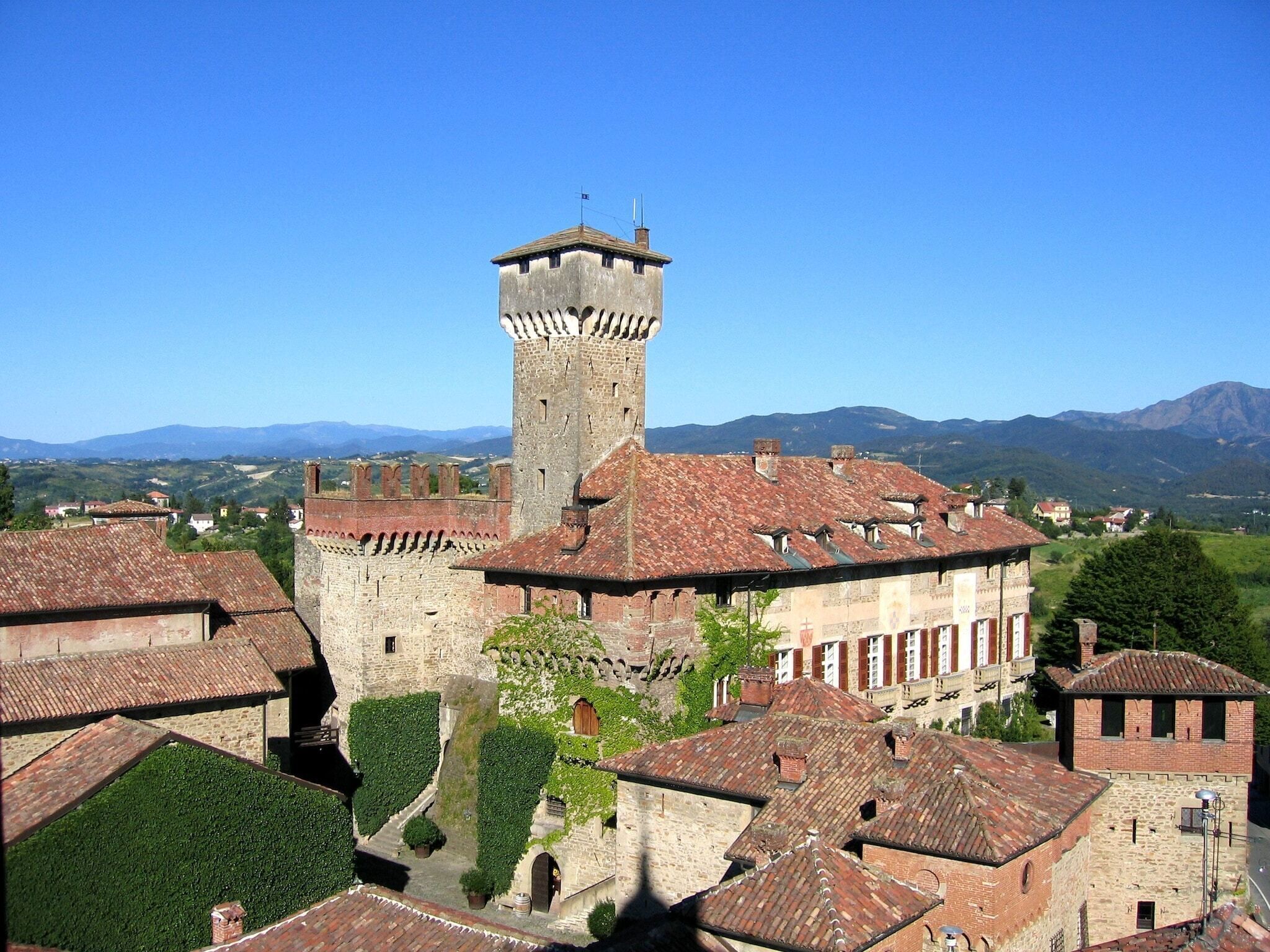 Un bâtiment historique en pierre, doté d’une tour imposante, entouré de toits traditionnels recouverts de tuiles rouges et d’une végétation luxuriante.