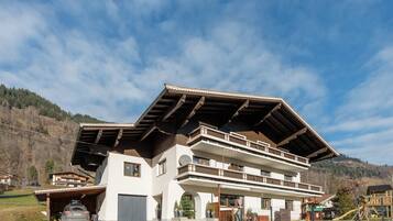 Chalet-style building with a sloped roof and balcony, set amidst a driveway and stone wall.