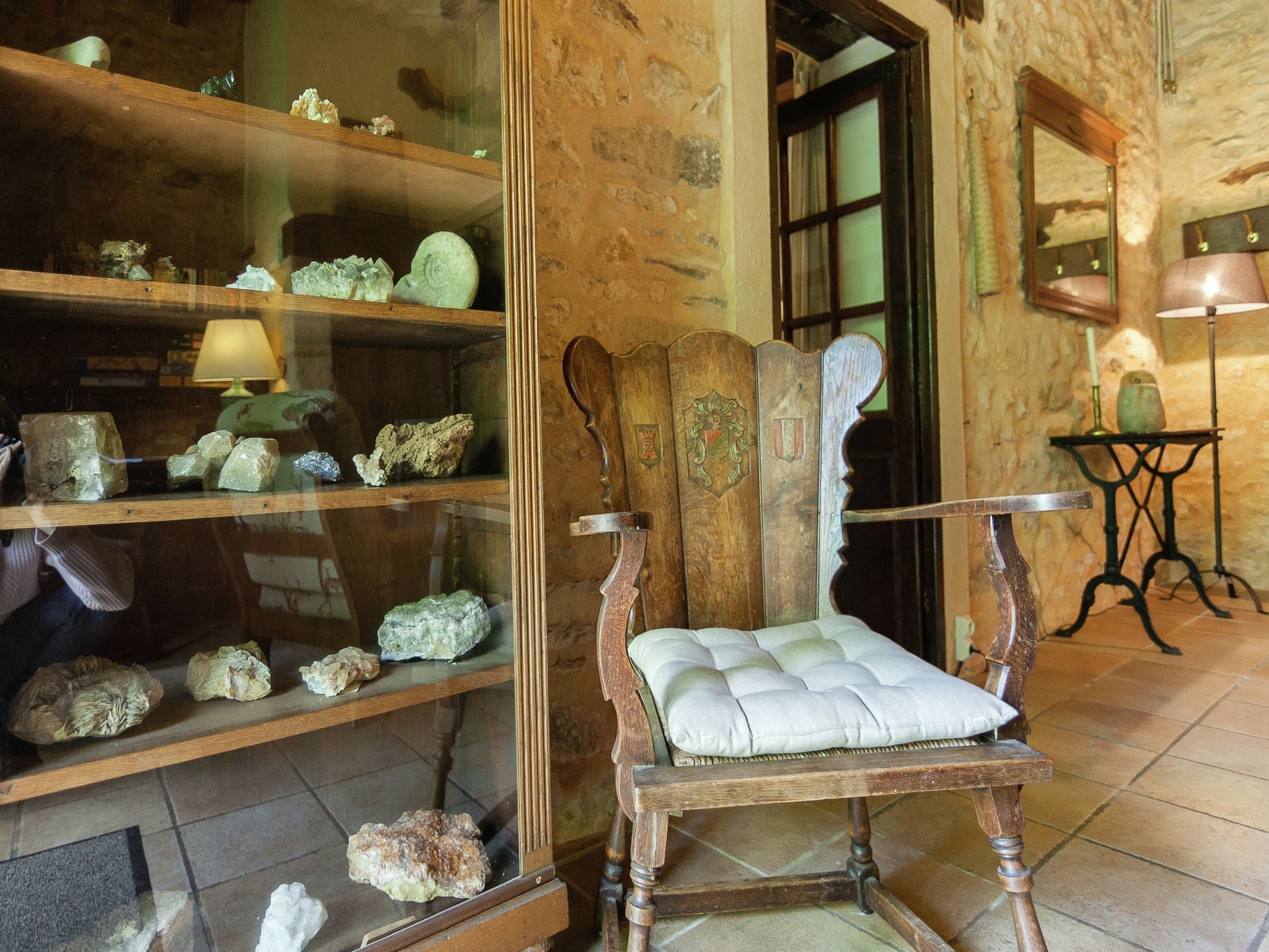 Un intérieur rustique avec une chaise en bois dotée d’un siège rembourré, une étagère en bois où sont exposés divers minéraux, et un miroir accroché au mur.