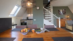 A dining area with a wooden table, black placemats, a bowl of fruit, and a magazine. A pendant light hangs above.