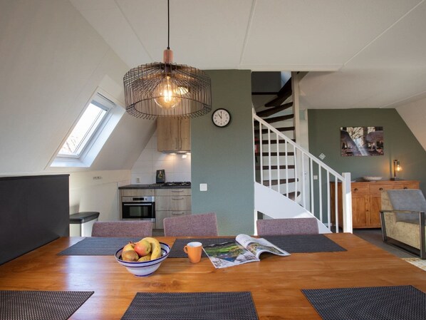 A dining area with a wooden table, black placemats, a bowl of fruit, and a magazine. A pendant light hangs above.