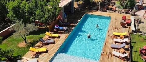 A rectangular pool with a wooden deck, surrounded by lounge chairs and umbrellas, set in a lush, green environment.