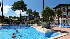 A swimming pool with two people swimming, surrounded by lounge chairs and palm trees. - Villa in France by Golf Course with Pool (Gujan Mestras)