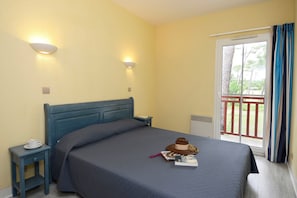 A single bed with a headboard, a bedside table with a cup, a hat, and a book on the bed. There are two wall-mounted lights and a window with curtains.