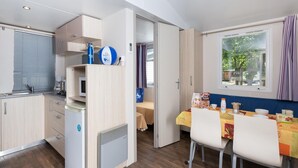 Cocina compacta con microondas integrado, cafetera y dispensador de agua. Hay una zona de comedor con una mesa preparada para dos personas y una ventana con cortinas.