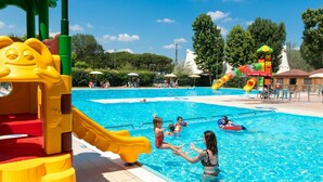 Una piscina con tobogán y zona de juegos acuáticos, rodeada de árboles y sombrillas.