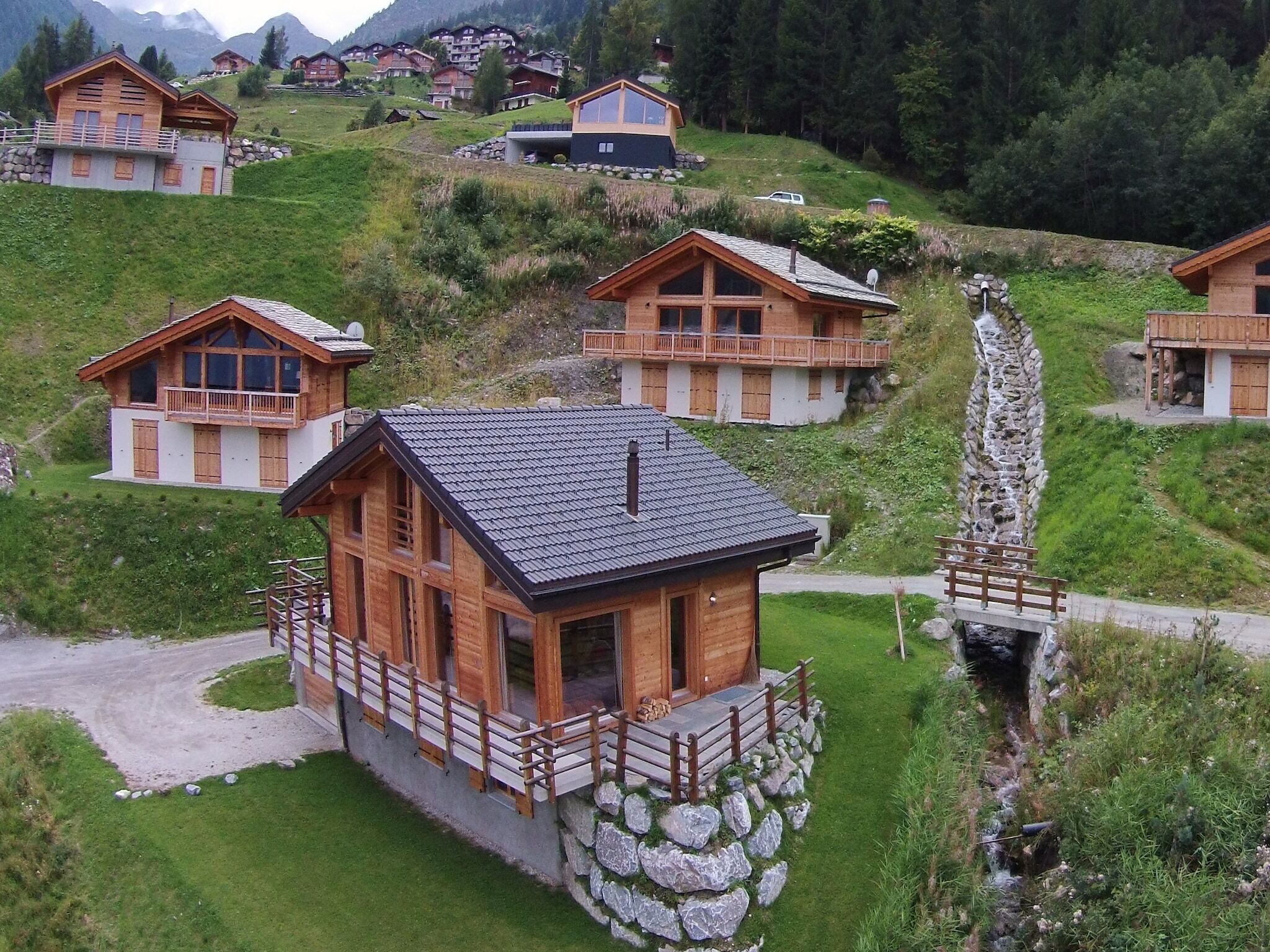 A cluster of wooden chalets on a hillside with stone foundations, surrounded by greenery and mountains.