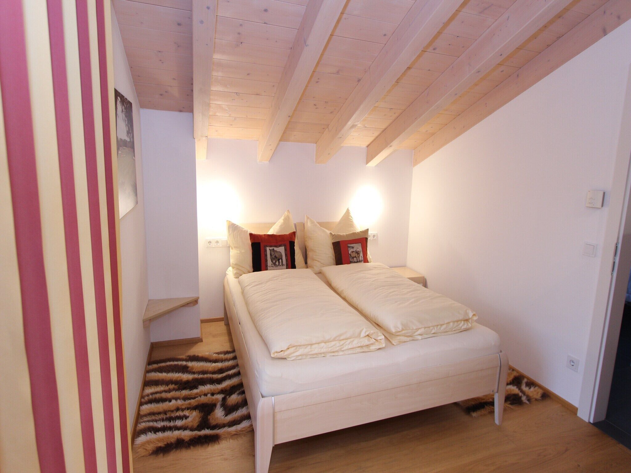 A bedroom with a wooden ceiling, a bed with two pillows, a small nightstand, and a fur rug.