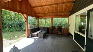 Uma área de varanda coberta com um banco, cadeiras, uma mesa pequena e vista para o verde ao redor.