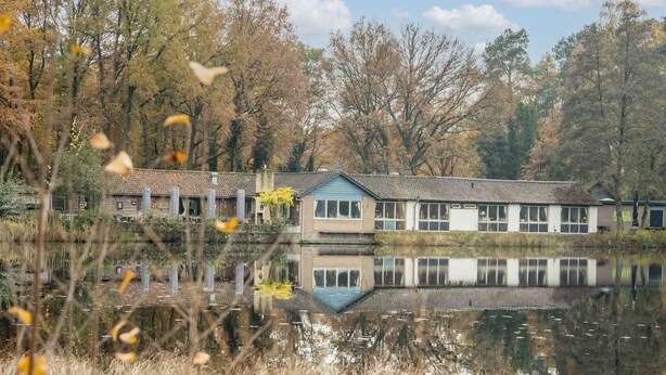 Um edifício à beira do lago com seu reflexo na água, rodeado por árvores e vegetação.