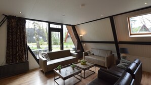 Un salon moderne comprenant un canapé, deux fauteuils, une table basse et une lampe. Une grande fenêtre, agrémentée de rideaux, offre une vue sur l’extérieur. Le plafond est en pente et les poutres apparentes ajoutent au charme de la pièce.