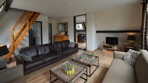 Un salon moderne avec un canapé en cuir marron, un canapé gris, une table basse en bois agrémentée de plantes, un meuble pour la télévision et un escalier.