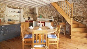 A cozy kitchen with a stone wall, wooden ceiling beams, a wooden staircase, and a dining area with wooden chairs and a table. - Cottage in Dordogne with Accessible Terrace (Roussines)