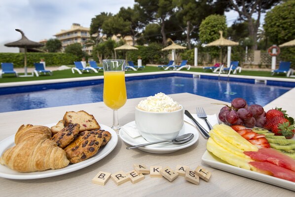 A breakfast setting with croissants, fruit, and a glass of orange juice by a poolside.