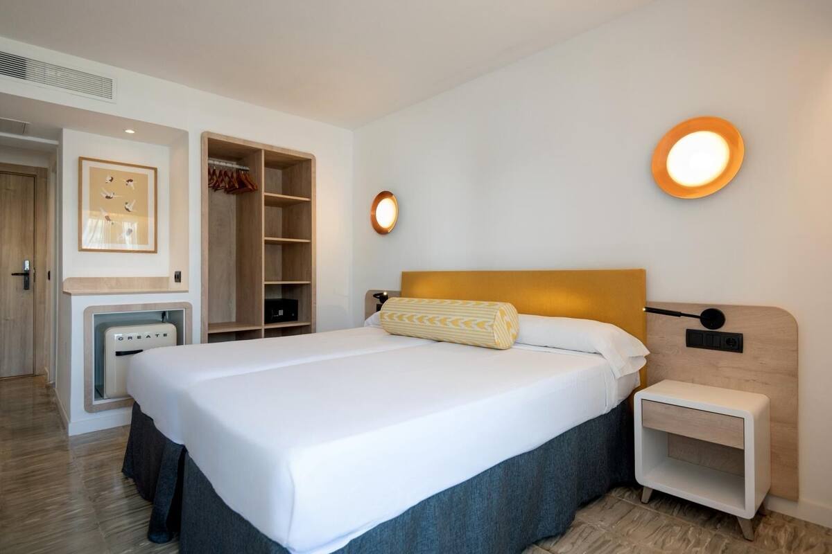 Quarto de hotel moderno com uma cama grande, mesa de cabeceira, guarda-roupa e luminária fixada na parede.