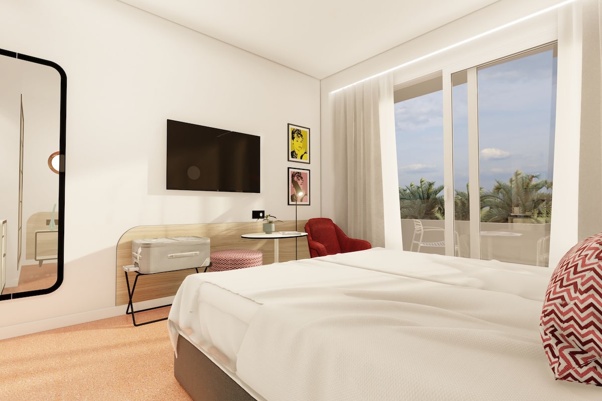 Quarto de hotel com cama, escrivaninha, cadeira, televisão e janela com vista para palmeiras.