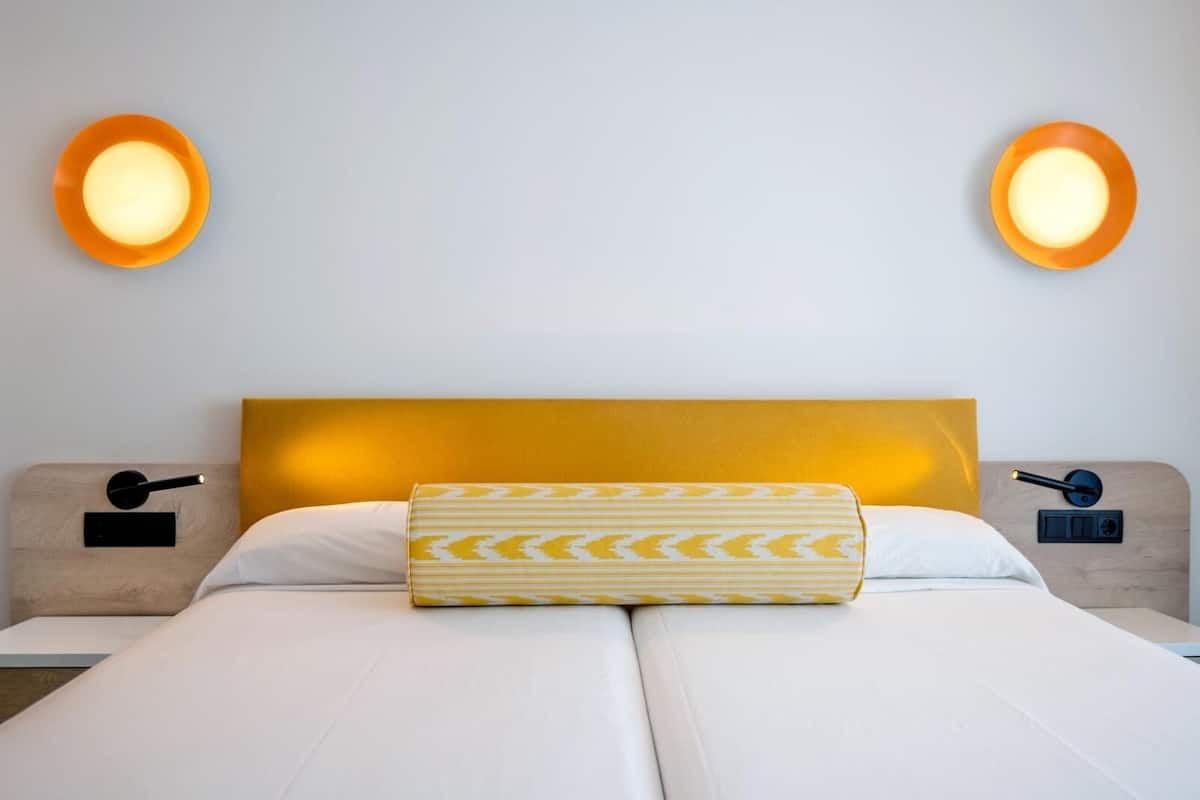 Uma cama de casal com cabeceira amarela, duas luminárias de apoio e almofada estampada.