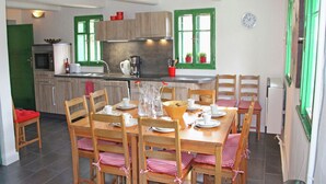 A dining area with a wooden table set for four, surrounded by chairs with checkered cushions. A kitchen with green windows and wooden cabinets is visible in the background.