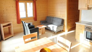 Een knus kamer in chaletstijl met houten inrichting, een grijze bank, een eettafel met stoelen en een kitchenette.