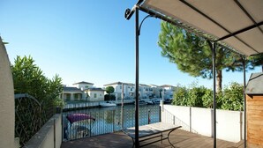 A wooden deck with a metal railing, a covered area with a canopy, and a view of a marina with boats and buildings in the background.