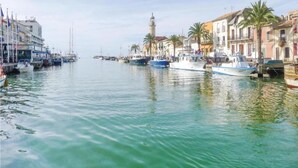 A calm waterfront with boats docked along the pier, surrounded by buildings with balconies and palm trees.