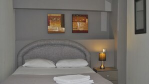 Ein ordentlich bezogenes Bett mit grauem Kopfteil, einer Nachttischlampe und zwei gerahmten Kunstwerken an der Wand.