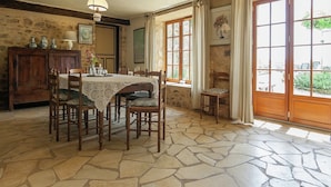 A stone-floored dining area with a table set for four, wooden chairs, a hanging light fixture, and a view through large windows. - Country House in Dordogne near Lascaux Cave (Les Eyzies-De-Tayac-Sireuil)