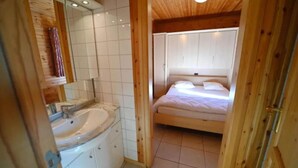 A bedroom with a wooden ceiling, a bed with white bedding, and a bathroom with a sink and mirror. - Holiday Home in La Roche with Sauna & Views (La Roche)