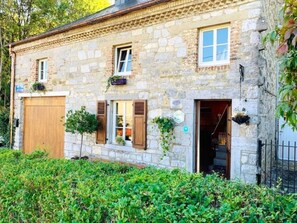 Un bâtiment en pierre avec une porte en bois, des fenêtres avec des volets et un petit arbre devant.