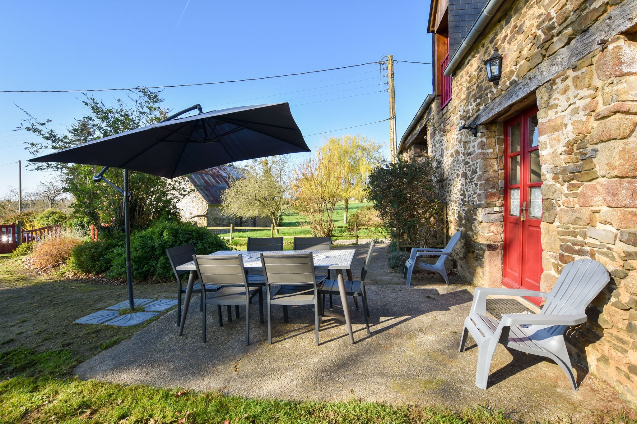 An outdoor patio with a table and chairs, a large umbrella, and a stone building with a red door.