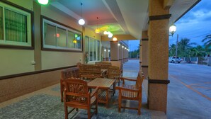 A covered outdoor seating area with wooden tables and chairs, a tiled floor, and a view of the street with palm trees and parked cars. - V Residence (Nakhon Ratchasima)