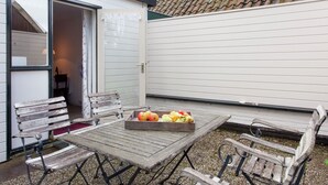 Une terrasse extérieure avec une table et des bancs en bois, un plateau de fruits sur la table et, en arrière-plan, un bâtiment avec un toit recouvert de tuiles.