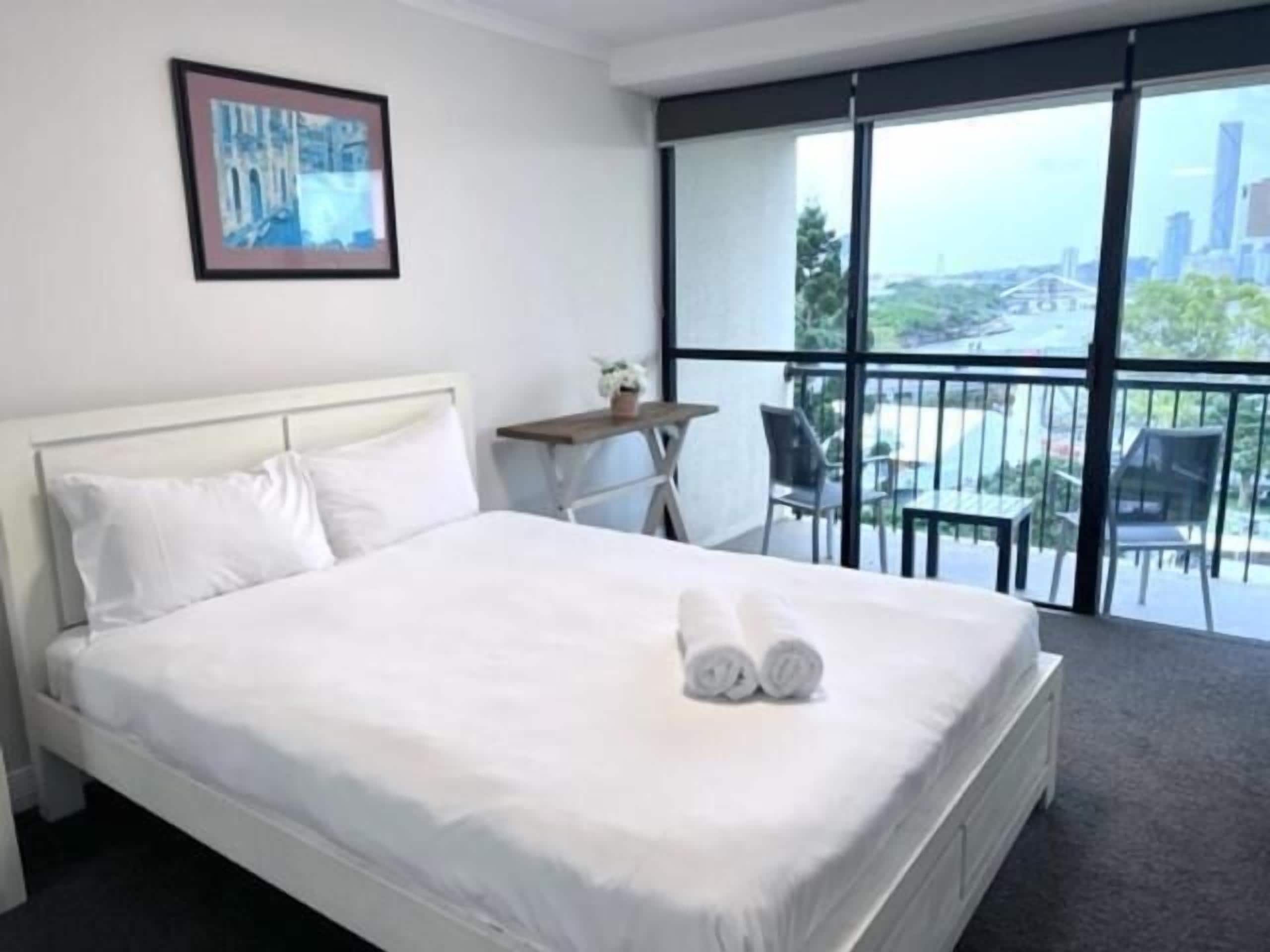 Habitación de hotel con cama, una mesita con una silla, balcón con vista a la ciudad y un cuadro en la pared.