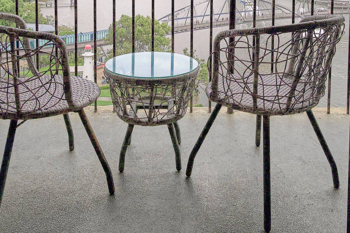 A balcony with two wicker chairs and a glass table, overlooking a river and city skyline.