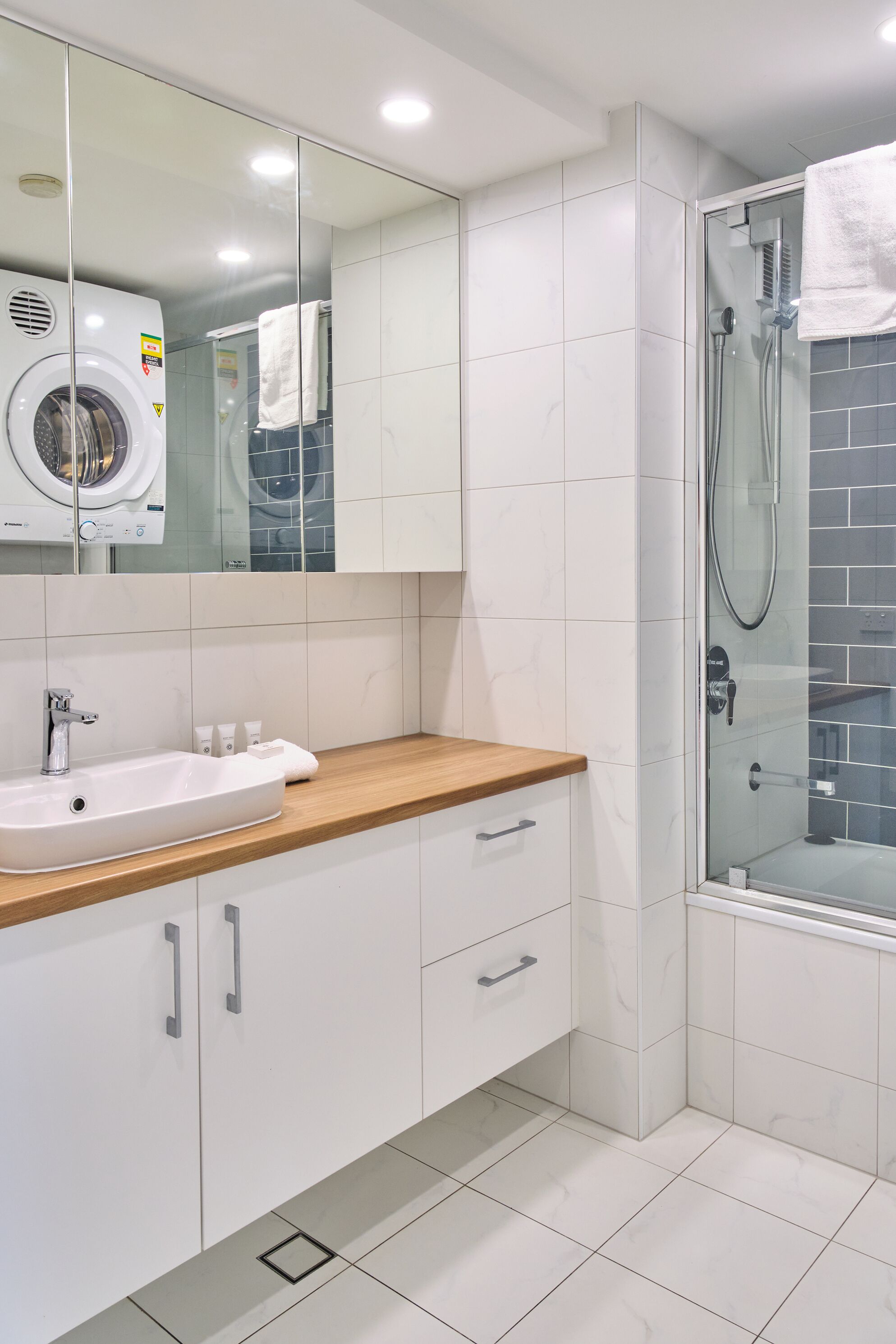 A modern bathroom with a built-in washing machine, a sink with a wooden countertop, and a glass-enclosed shower area.